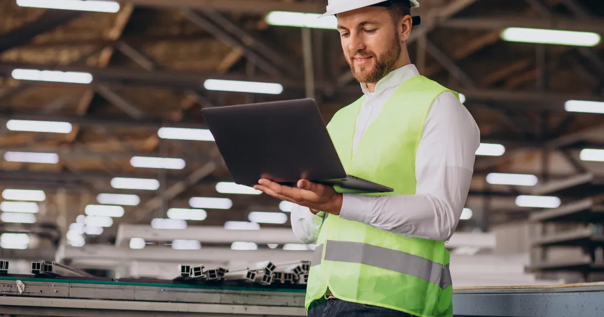 Engenheiro de capacete branco e colete refletivo segurando um notebook aberto dentro de um galpão industrial