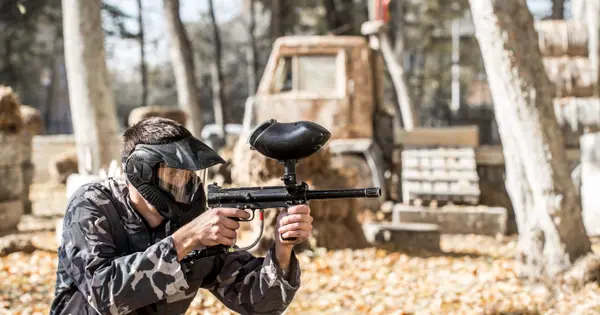 Jogador de paintball com uniforme camuflado e máscara, agachado e mirando o marcador num campo ao ar livre com obstáculos e folhas secas no chão