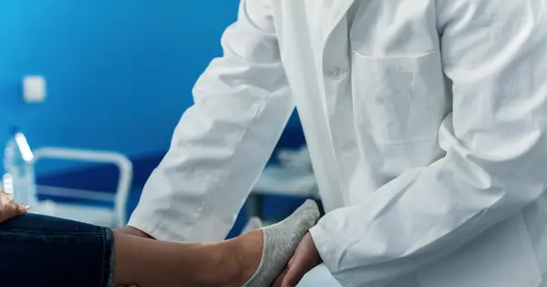 Profissional de jaleco branco segurando e examinando o pé de uma paciente sentada na maca de um consultório
