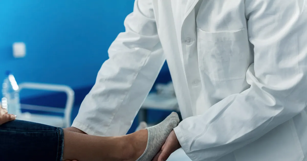 Profissional de jaleco branco segurando e examinando o pé de uma paciente sentada na maca de um consultório