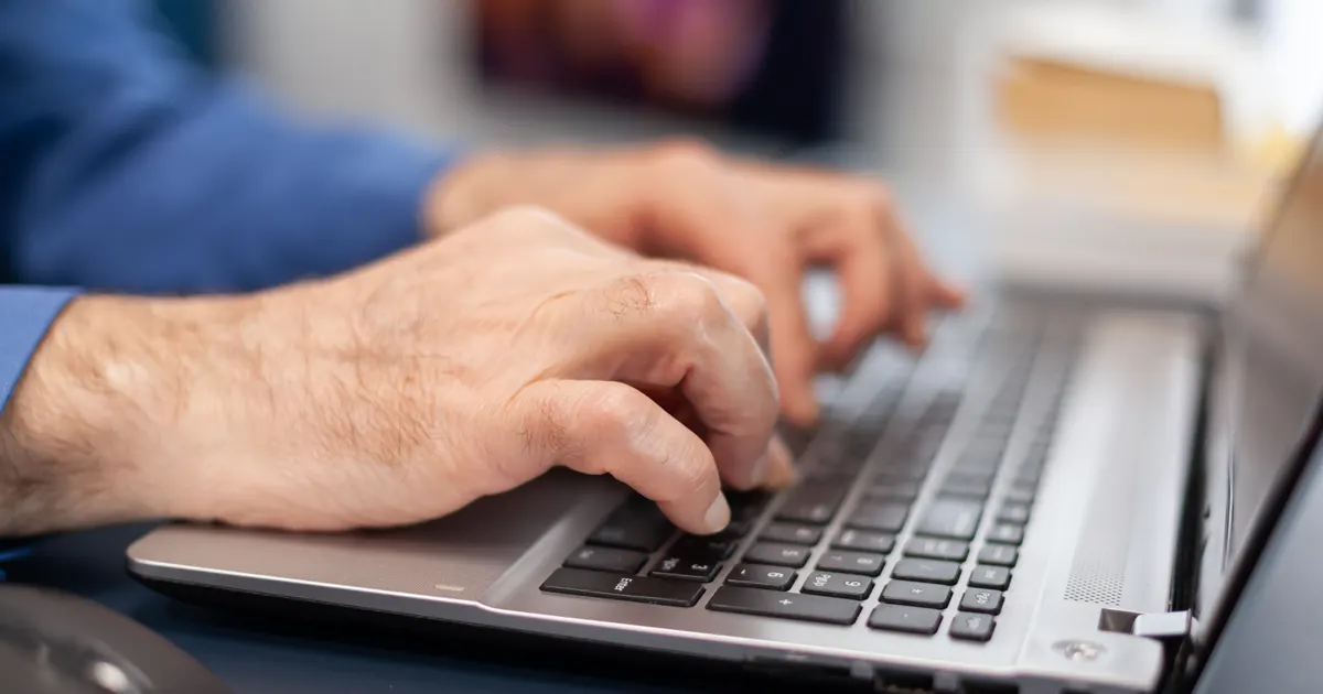 Mãos de um homem mais velho digitando no teclado de um notebook prateado, com um mouse ao lado sobre a mesa