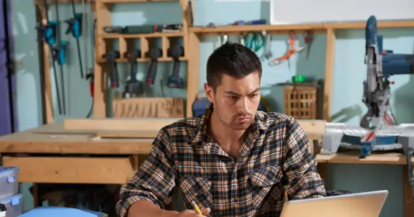 Homem de camisa xadrez sentado a uma mesa de madeira numa oficina de marcenaria, olhando para um notebook e anotando num papel, com ferramentas penduradas na parede ao fundo