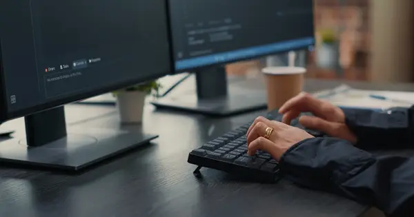Mãos de programador digitando código no teclado diante de dois monitores