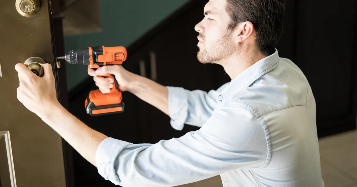 Homem de camisa clara usando uma parafusadeira laranja para fixar a maçaneta de uma porta de madeira, com uma escada ao fundo